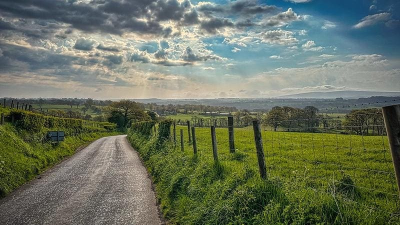 A narrow country road winds through lush green fields, bordered by a rustic fence and hedgerows. The scene is bathed in warm sunlight filtering through a dramatic sky filled with clouds, casting soft rays over the distant rolling hills. The rural landscape stretches into the horizon, dotted with trees and farmland, creating a serene and picturesque countryside view. Photo was taken by the author on his 84 mile hike along Hadrian's Wall in northern England.